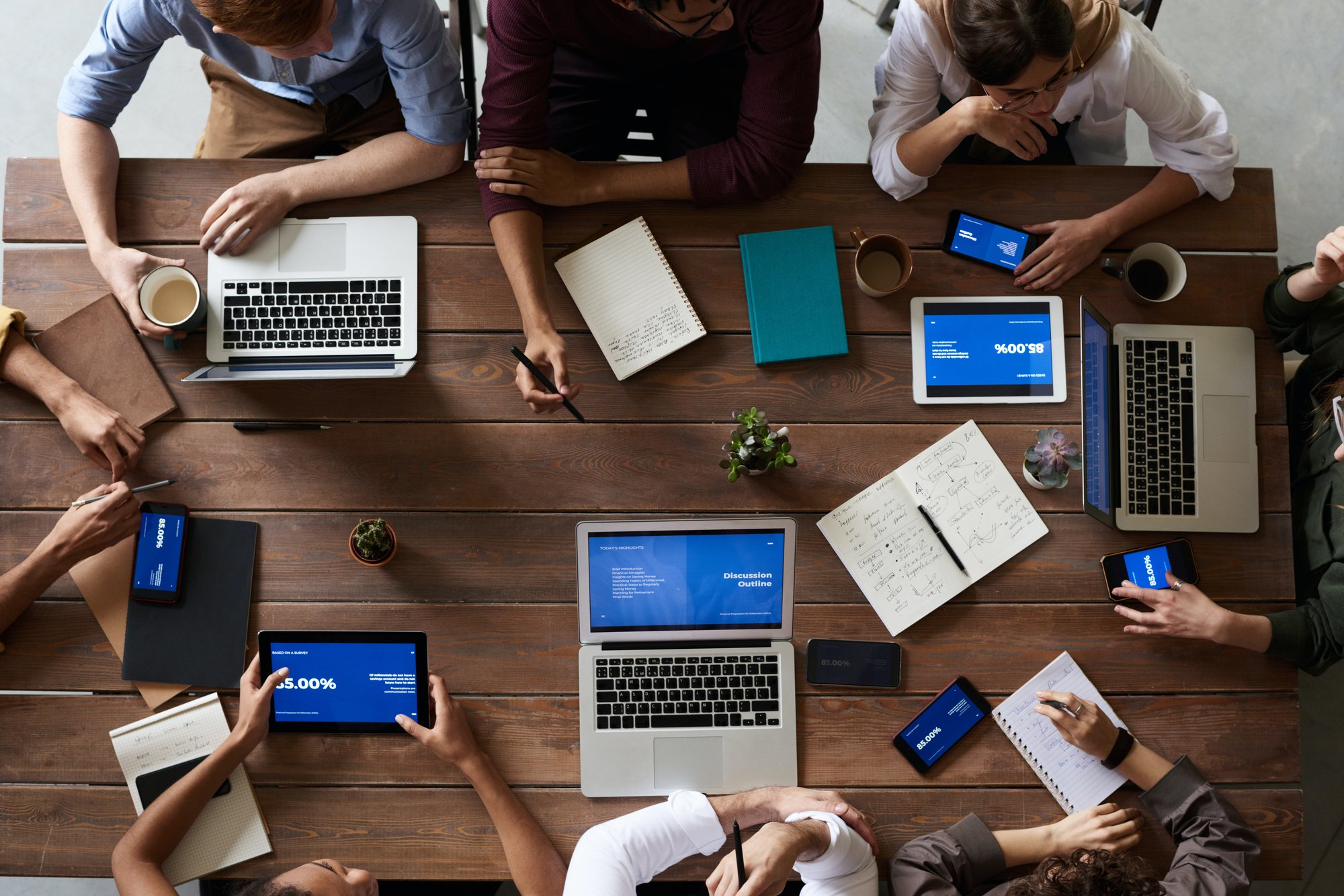 A group of coworkers sitting on a wooden table with their laptops, tables, phones, and notebooks on top.